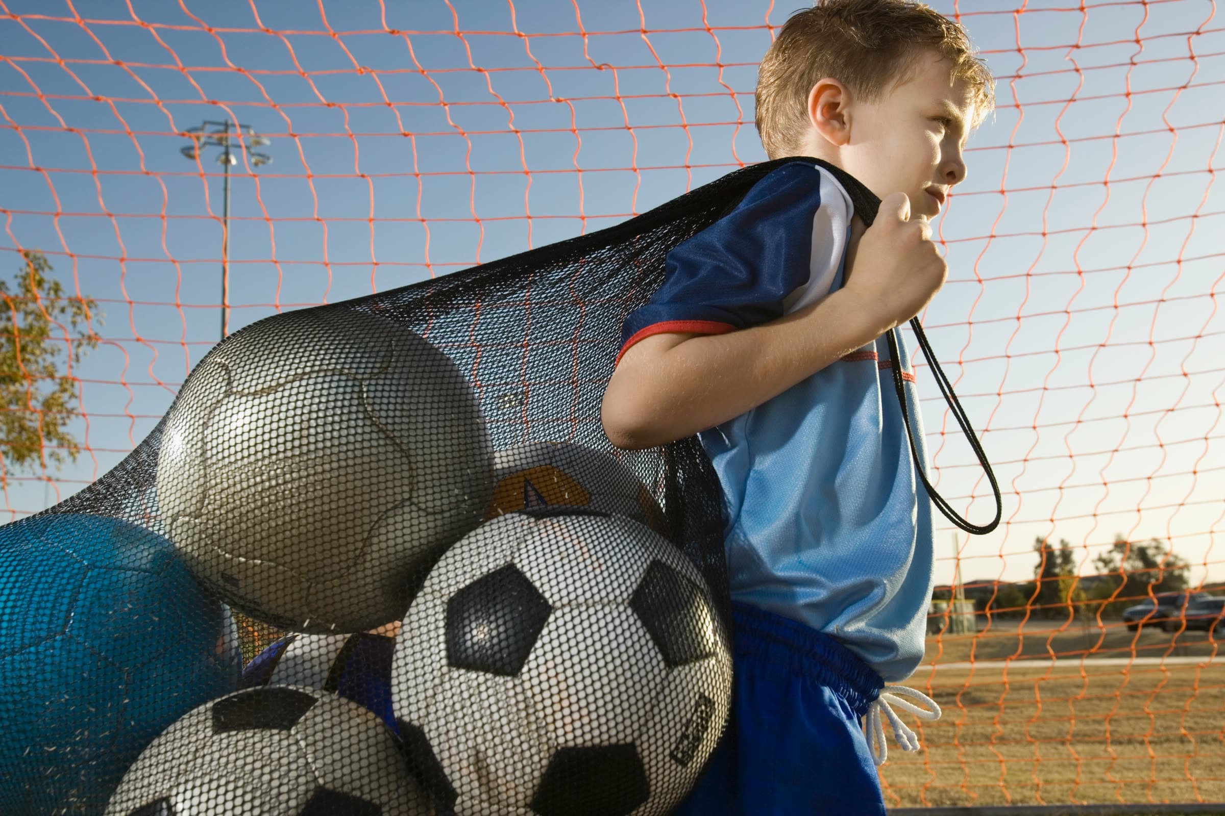 A boy carries a net full of footballs over his shoulder at sunset