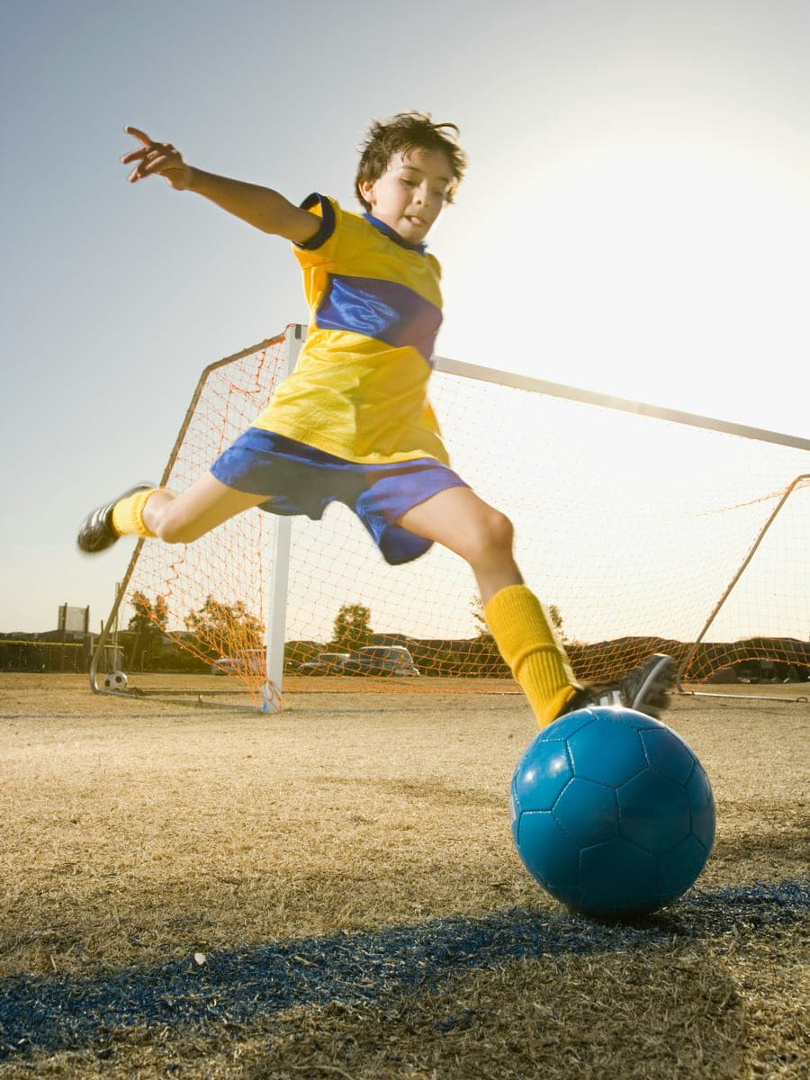 A young boy in a yellow kit swings his leg to kick a blue ball in front of a goal
