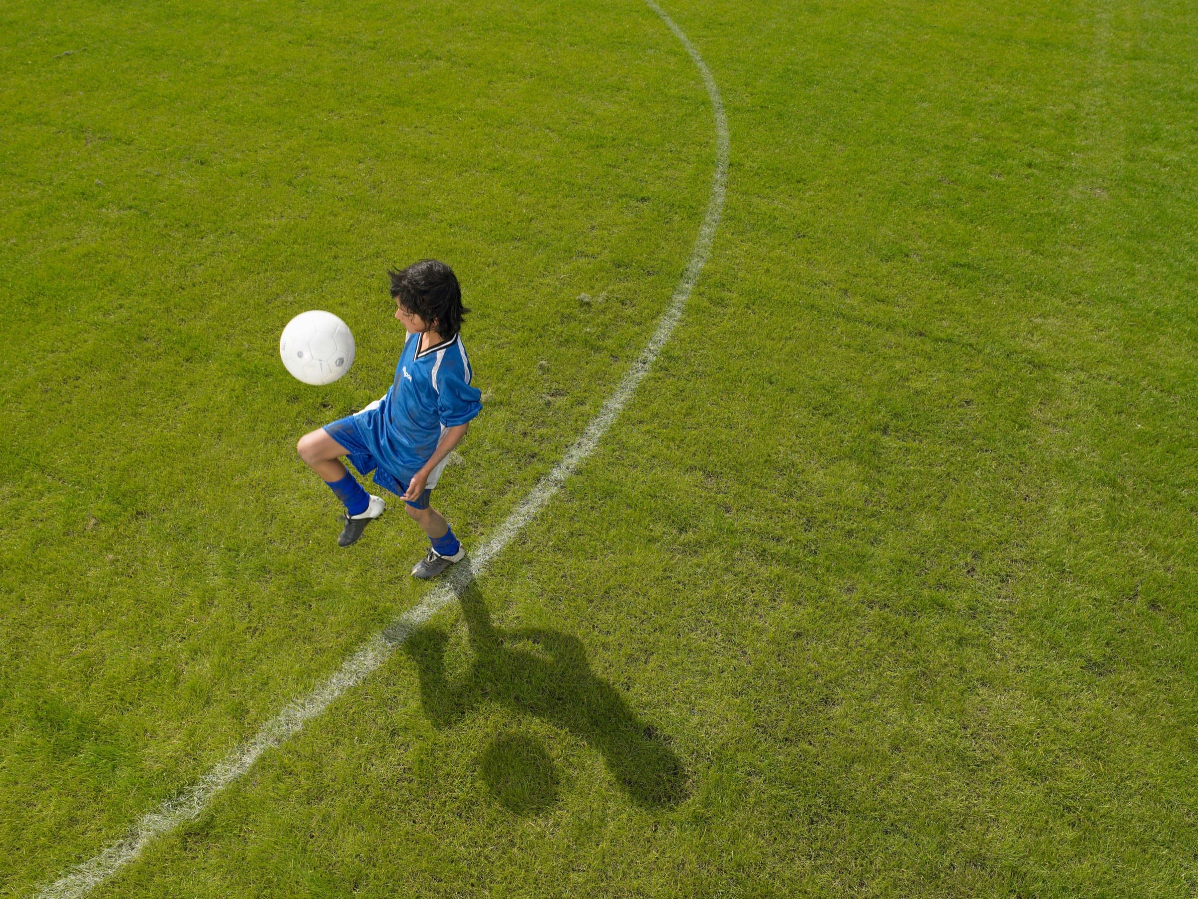 A boy in a blue kit controls the ball, photographed from above; his shadow falls long across the grass.